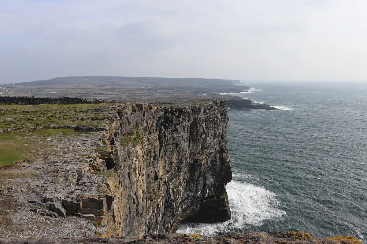 A sheer limestone sea cliff on Inis Mór with waves breaking at its base, grass and limestone pavement on top, and the low island stretching away under a hazy sky
